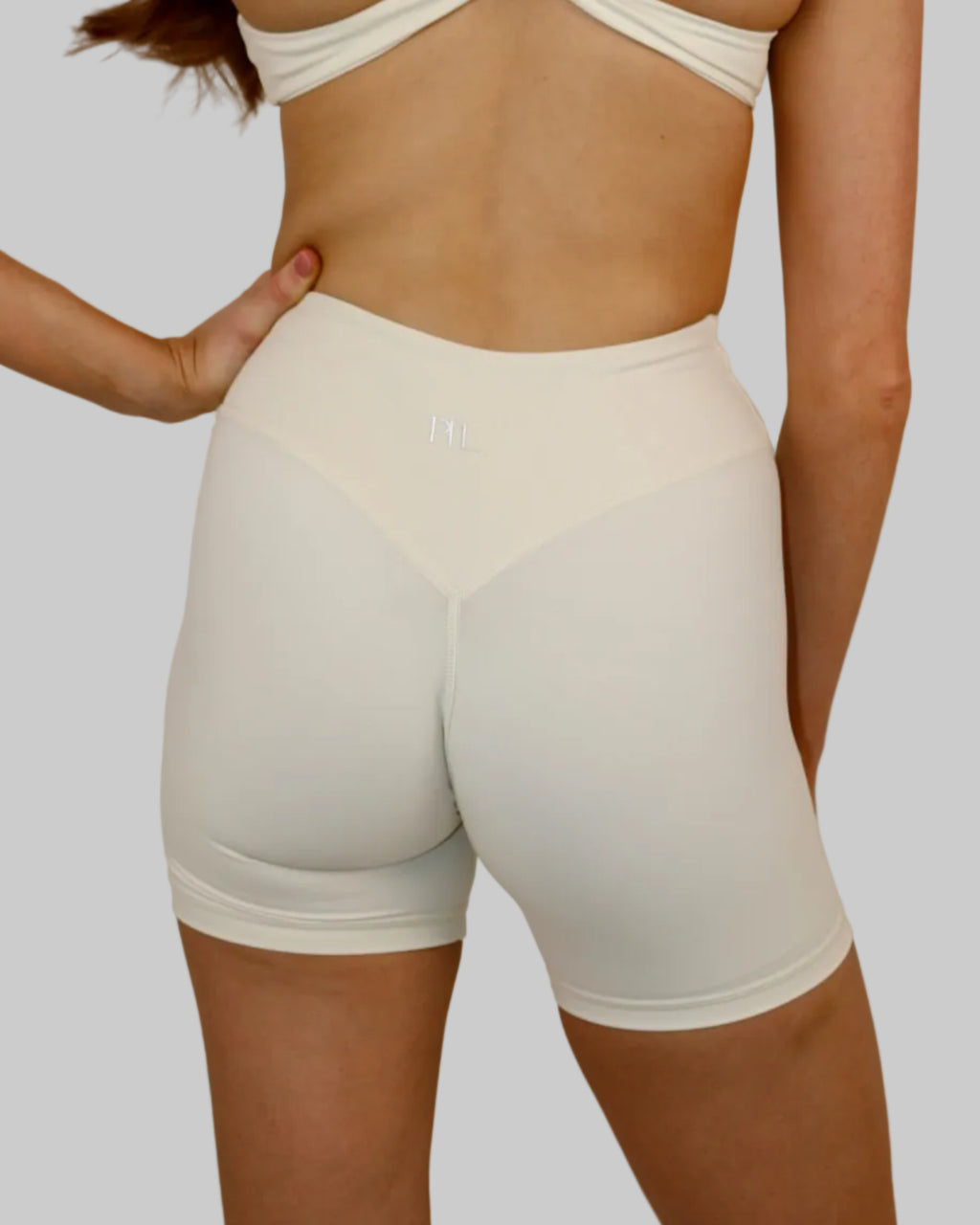 Rear view of a woman wearing white high-waisted athletic shorts and a matching white strappy sports bra against a plain light beige wall.