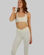 A woman modeling a matching off-white activewear set consisting of a sports bra and high-waisted leggings, photographed against a plain light gray studio background.