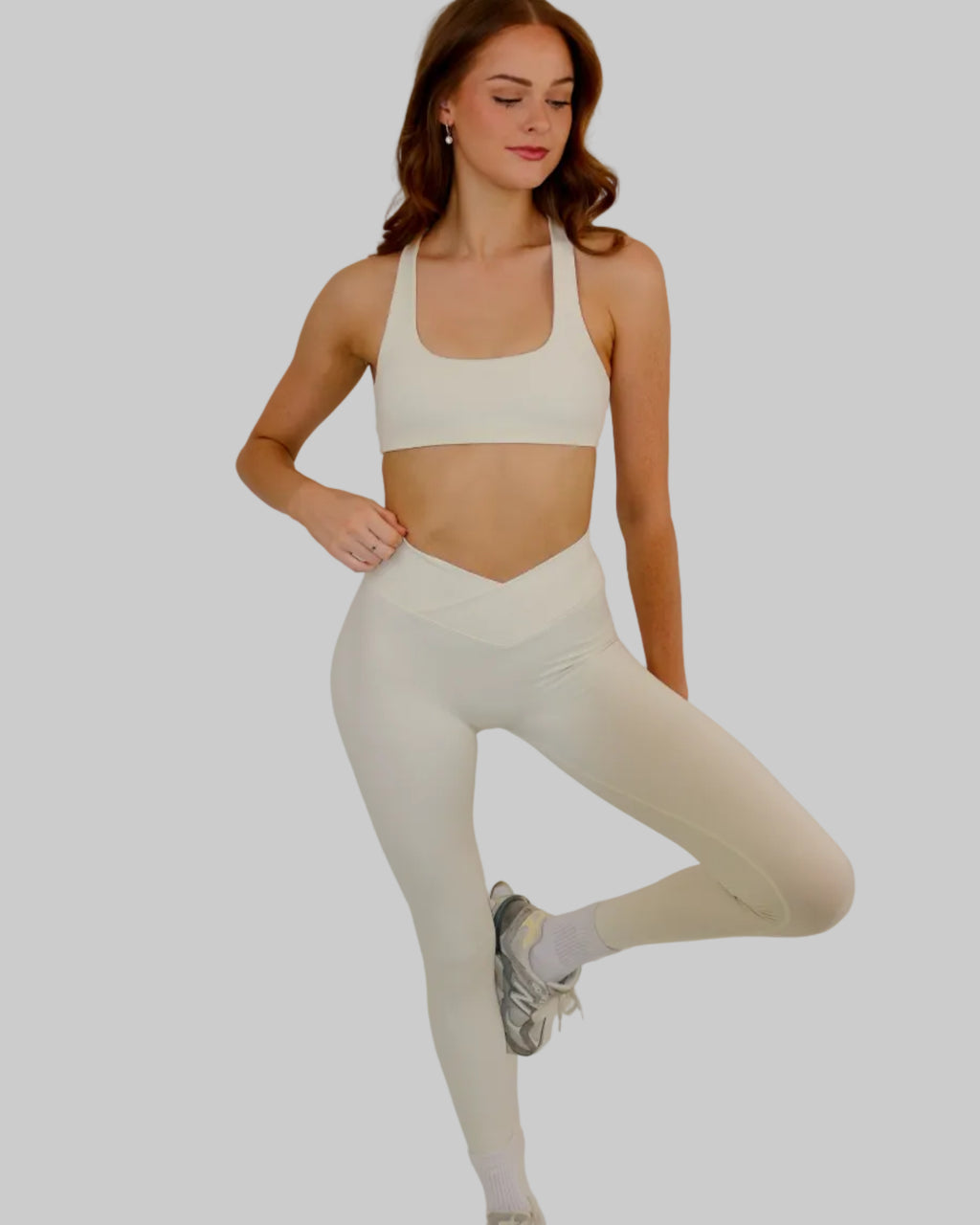 A woman modeling a matching off-white activewear set consisting of a sports bra and high-waisted leggings, posing against a plain light beige studio background.