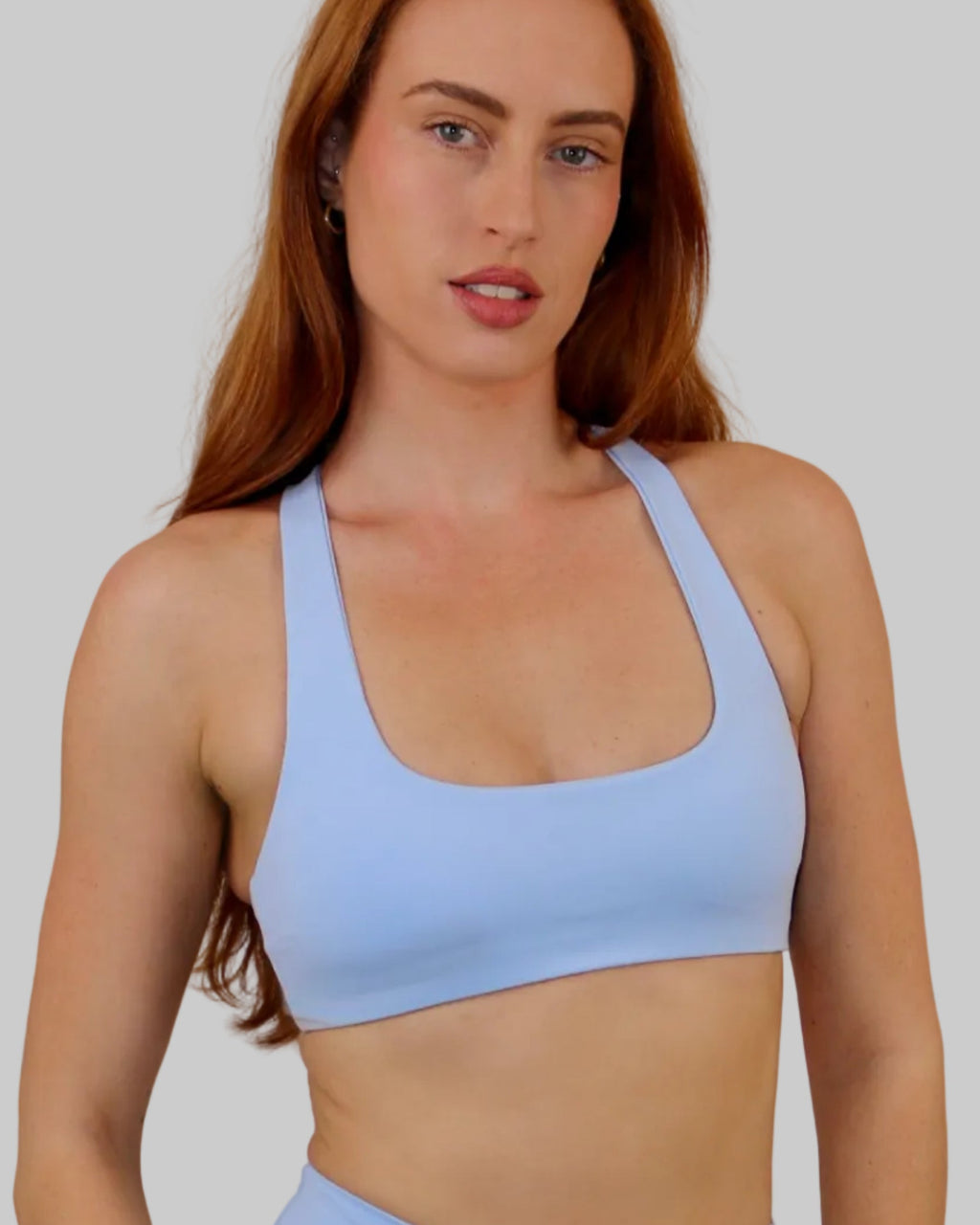 A woman modeling a light blue athletic scoop-neck sports bra and matching high-waisted shorts against a plain off-white wall.