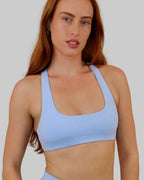A woman modeling a light blue athletic scoop-neck sports bra and matching high-waisted shorts against a plain off-white wall.