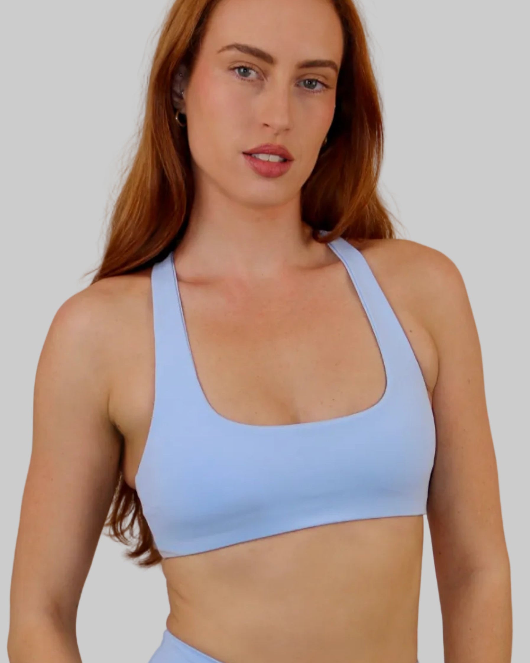 A woman modeling a light blue athletic scoop-neck sports bra and matching high-waisted shorts against a plain off-white wall.