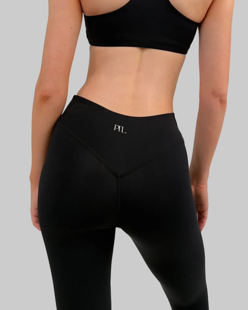 Rear view of a model wearing a black sports bra and matching high-waisted leggings with a small white 'FYL' logo on the back waistband, against a light gray studio background.