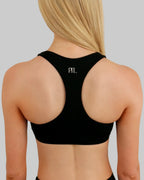 A woman viewed from the back wearing a black racer back sports bra with a small white 'PTL' logo centered on the upper back, against a plain light grey studio background.