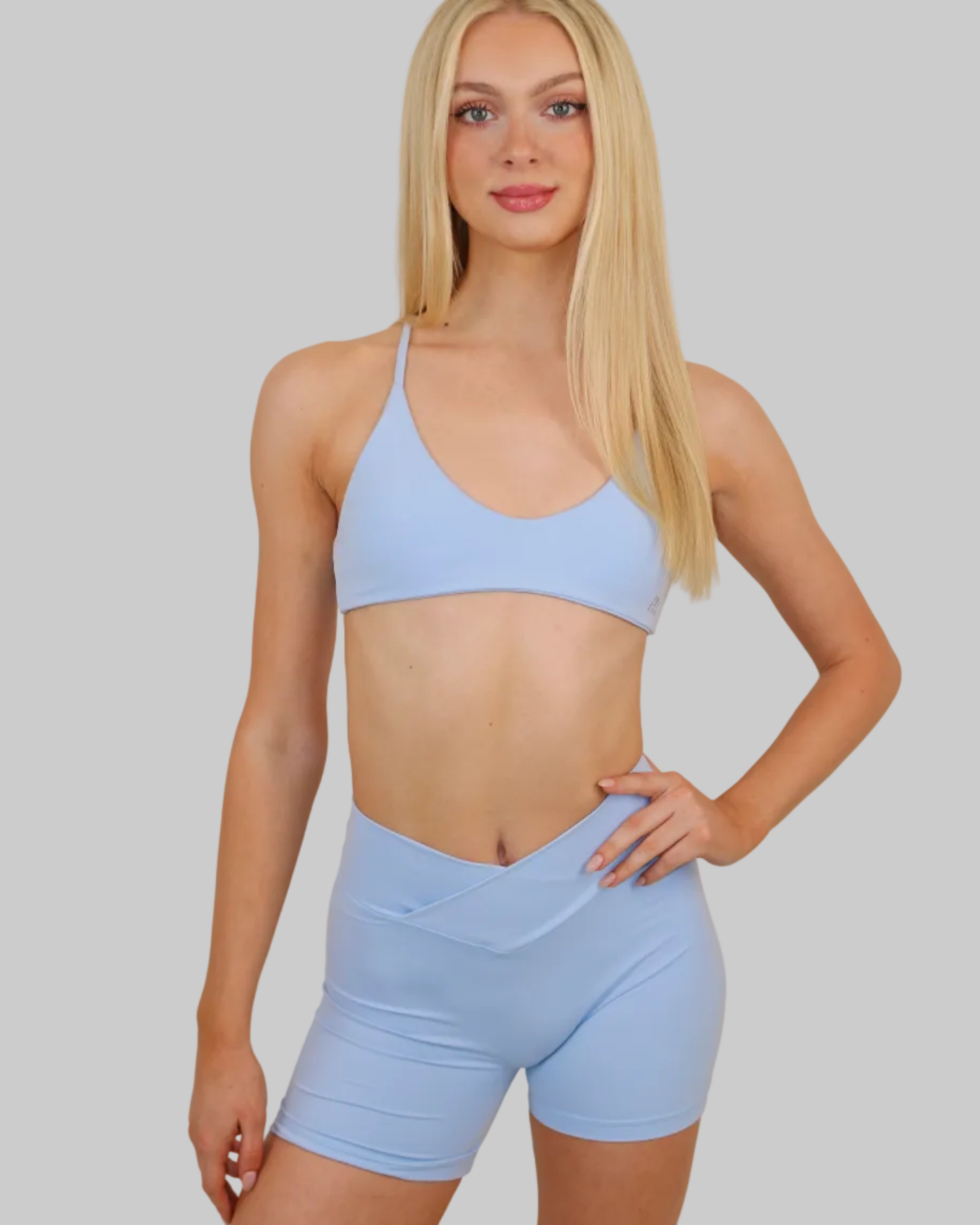A blonde model wearing a light blue two-piece activewear set consisting of a sports bra and matching high-waisted shorts, posing against a plain beige wall.