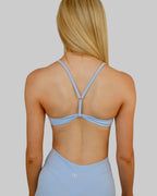 A woman viewed from behind wearing a light blue athletic two-piece set consisting of a sports bra and matching high-waisted shorts, set against a plain beige wall.