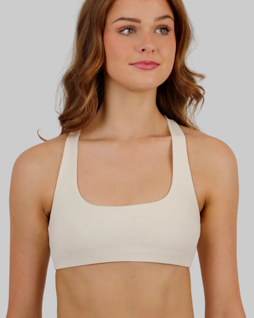 A woman with wavy red hair wearing a cream-colored square scoop-neck sports bra and matching high-waisted crop shorts, photographed against a solid light beige studio background.
