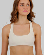 A woman with wavy red hair wearing a cream-colored square scoop-neck sports bra and matching high-waisted crop shorts, photographed against a solid light beige studio background.
