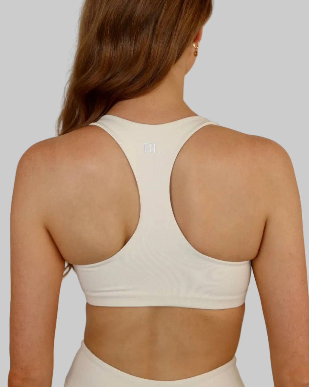 A woman viewed from the back wearing a white athletic racer back sports bra and matching bottoms against a plain light beige wall.