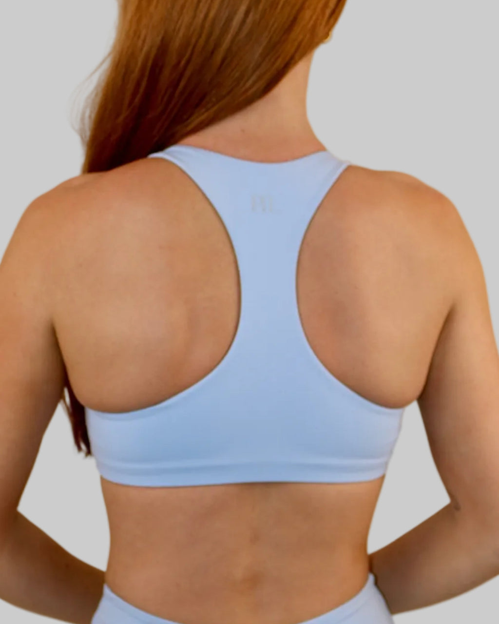 A woman with long red hair is shown from the back wearing a light blue sports bra and matching high-waisted athletic shorts against a plain beige wall.