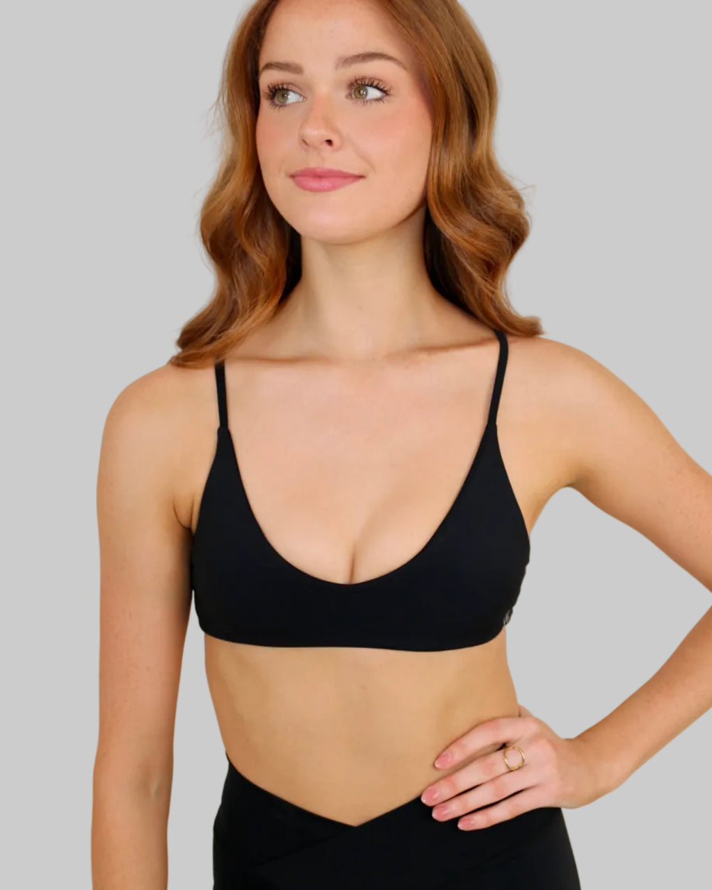 A woman with wavy red hair wearing a black scoop-neck sports bra and black high-waisted leggings, posing against a plain beige wall.