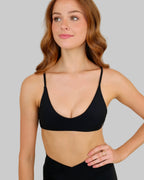 A woman with wavy red hair wearing a black scoop-neck sports bra and black high-waisted leggings, posing against a plain beige wall.