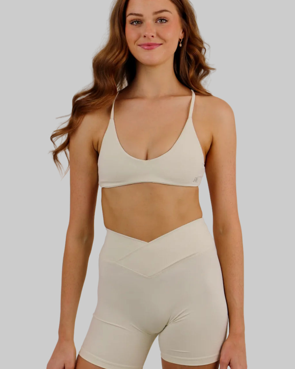 A woman modeling a matching off-white activewear set consisting of a sports bra and high-waisted shorts, set against a plain light beige studio background.
