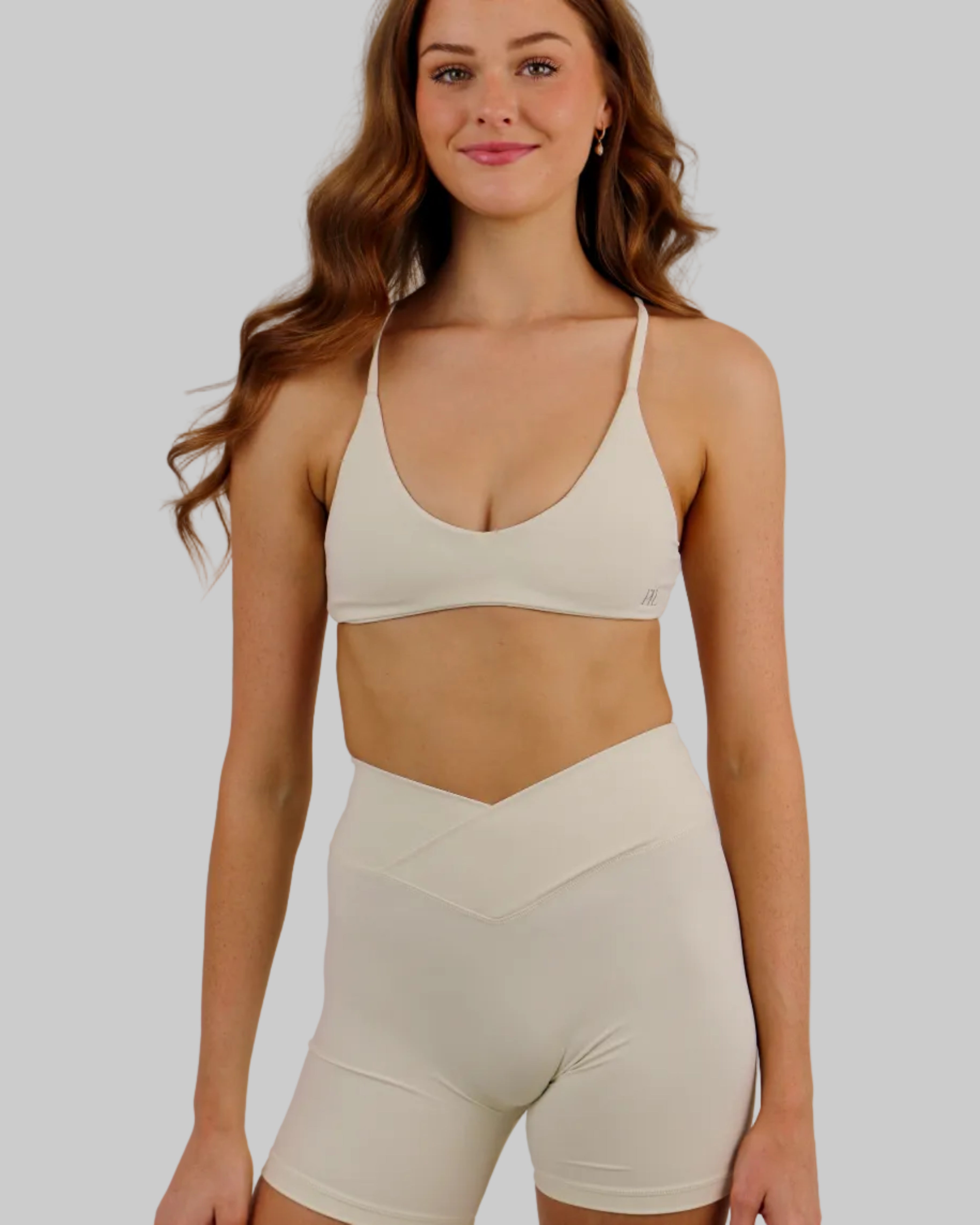 A woman modeling a matching off-white activewear set consisting of a sports bra and high-waisted shorts, set against a plain light beige studio background.