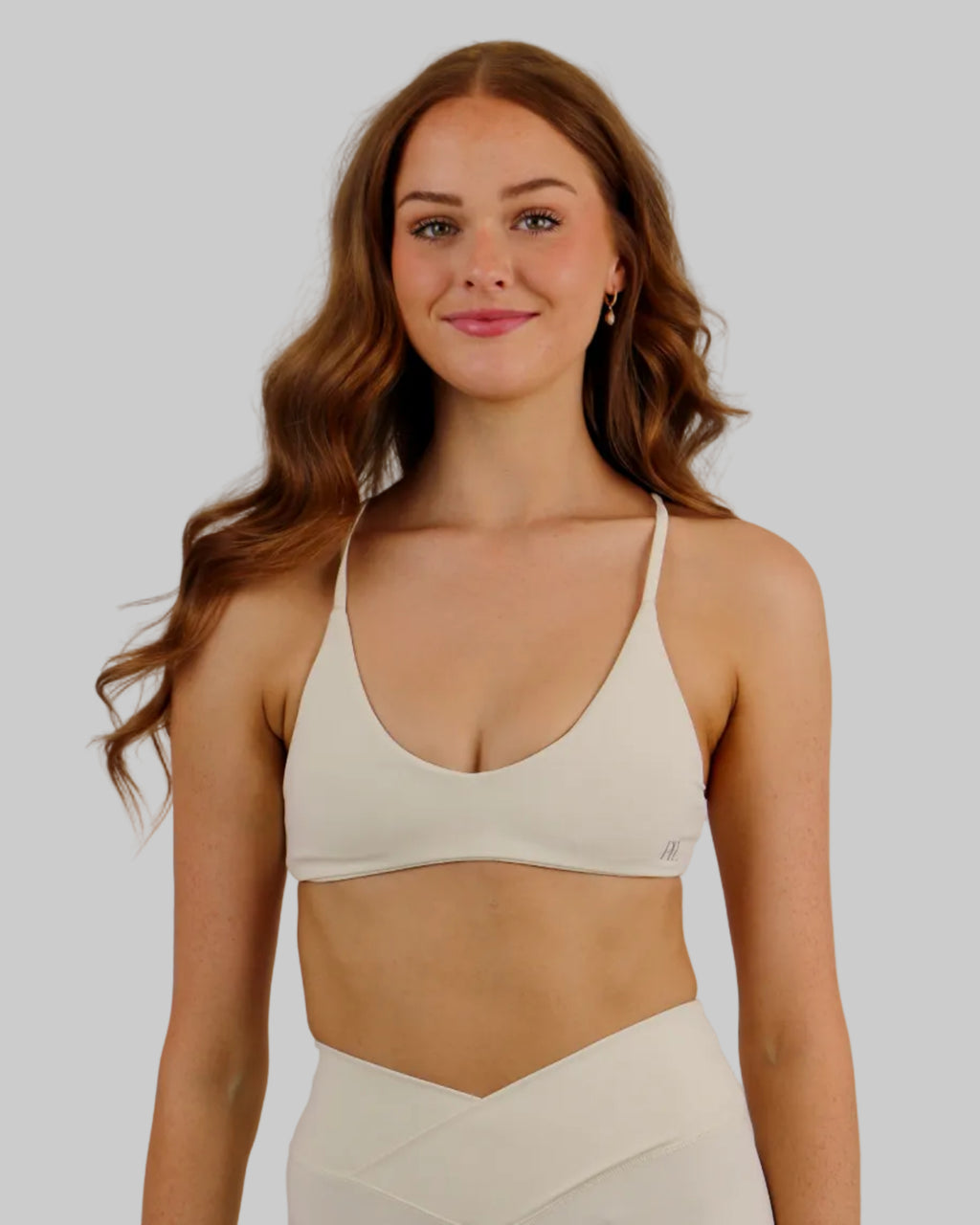 A woman modeling a matching off-white activewear set consisting of a sports bra and high-waisted shorts, set against a plain light beige studio background.