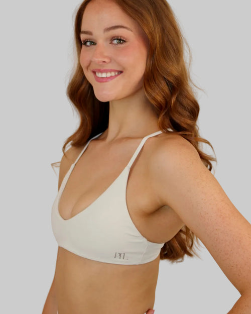A smiling woman with long wavy red hair wearing a white scoop-neck bralette with thin straps and a small 'PTL' logo on the lower band, posed against a plain light beige studio background.