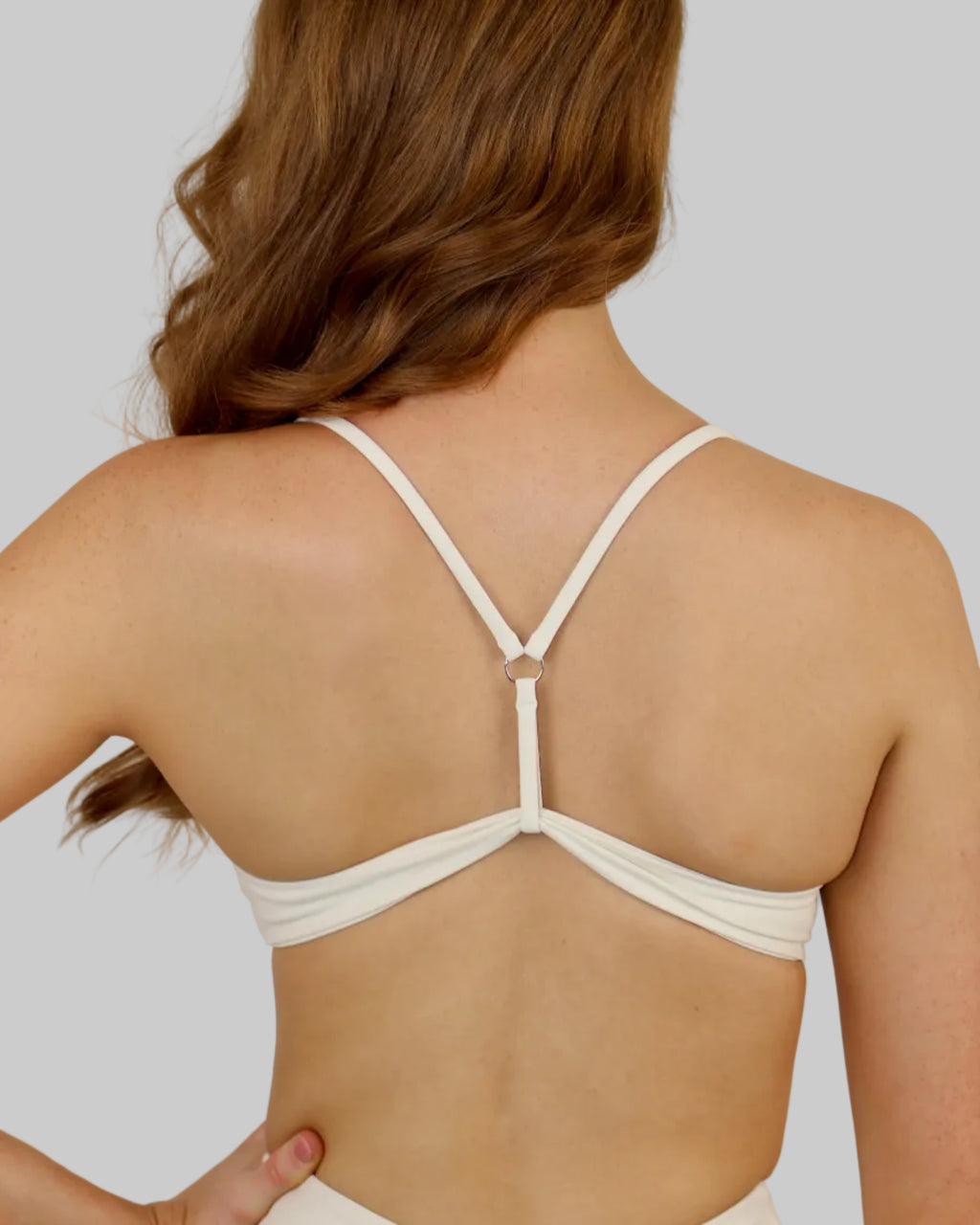 A woman shown from the back wearing a white strappy sports bra, with her hair styled in loose waves against a plain light beige background.