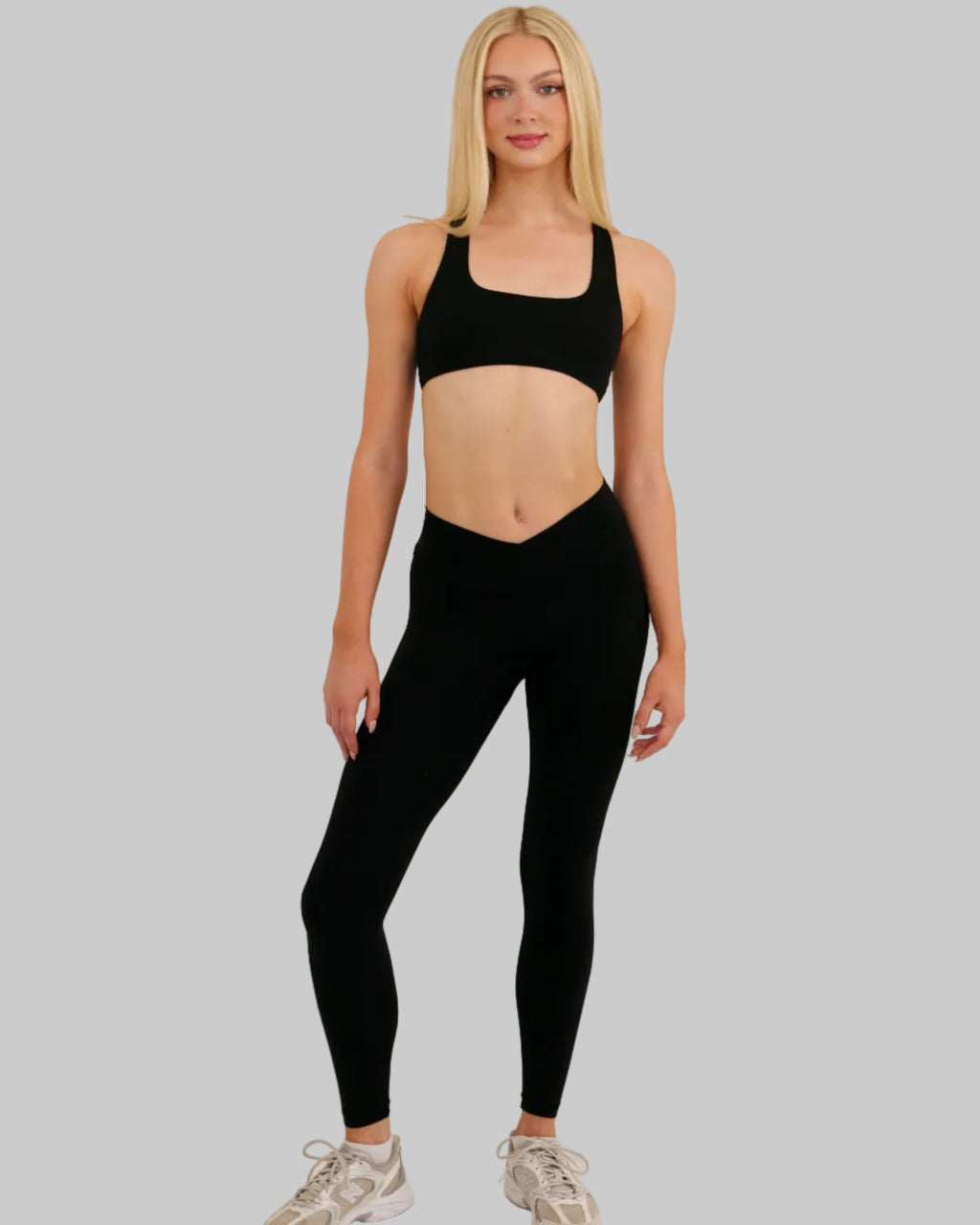 A blonde model stands against a plain light beige studio background wearing a black sports bra and matching black high-waisted leggings, paired with light beige athletic sneakers.