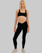A blonde model stands against a plain light beige studio background wearing a black sports bra and matching black high-waisted leggings, paired with light beige athletic sneakers.
