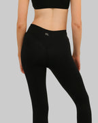 Rear view of a model wearing black high-waisted leggings with a small white 'PTL' logo on the back waistband, paired with a black sports bra, against a plain light beige background.