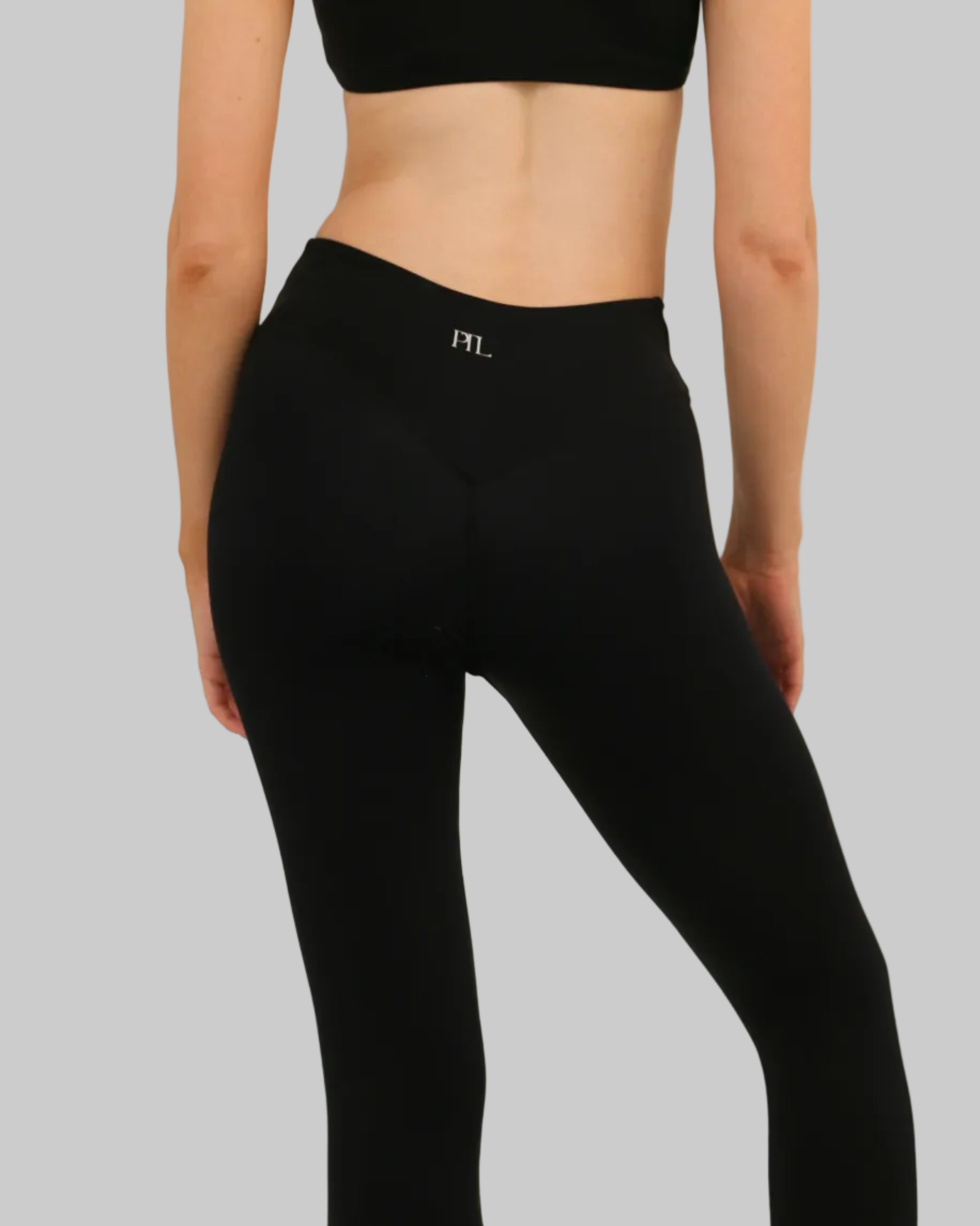 Rear view of a model wearing black high-waisted leggings with a small white 'PTL' logo on the back waistband, paired with a black sports bra, against a plain light beige background.