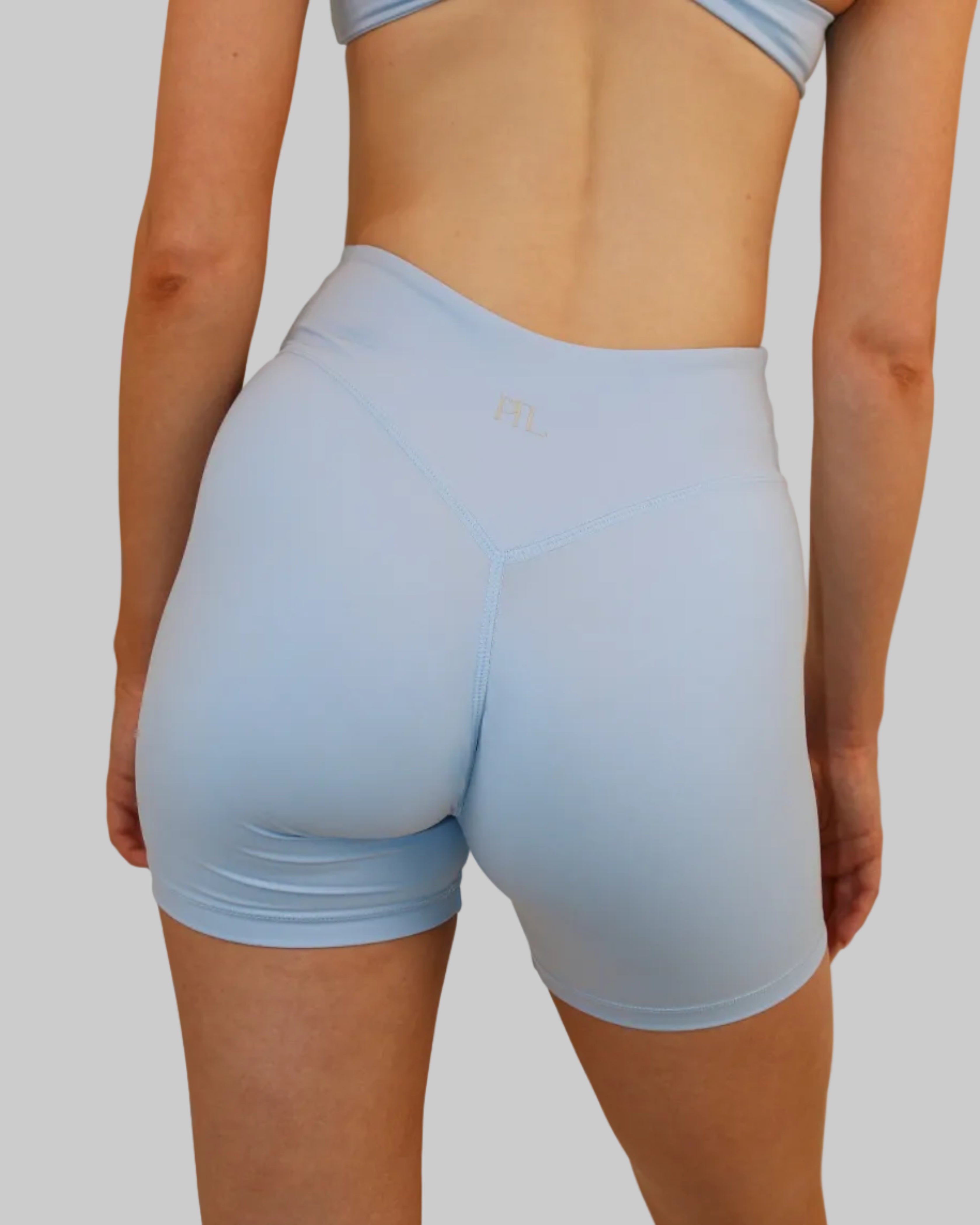 A person shown from the back wearing light blue athletic shorts and a matching light blue strappy top against a plain beige studio background.