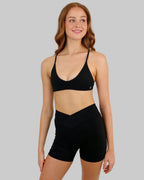 A woman modeling a matching black activewear set consisting of a scoop-neck sports bra and high-waisted biker shorts, set against a plain beige studio background.