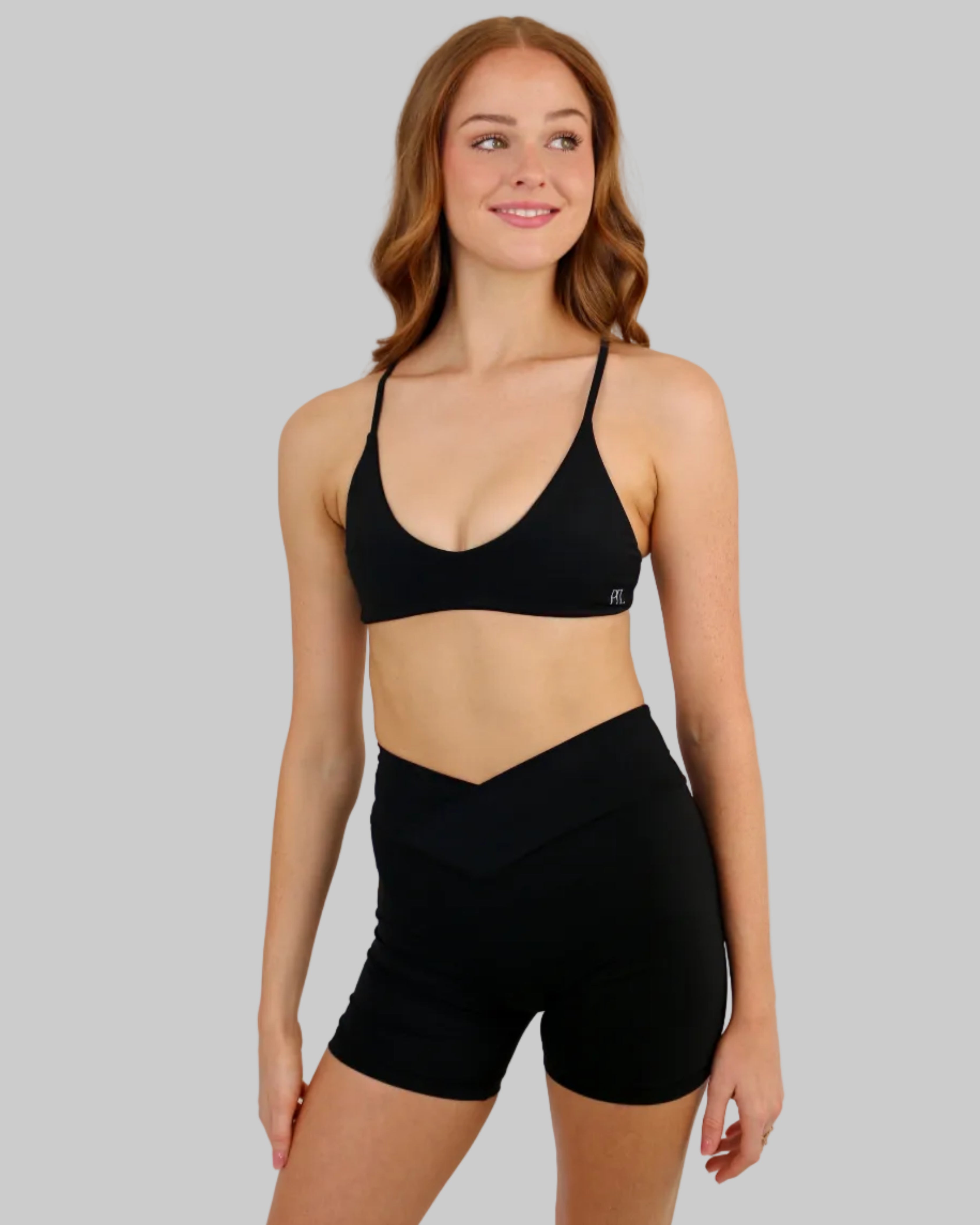 A woman modeling a matching black activewear set consisting of a scoop-neck sports bra and high-waisted biker shorts, set against a plain beige studio background.