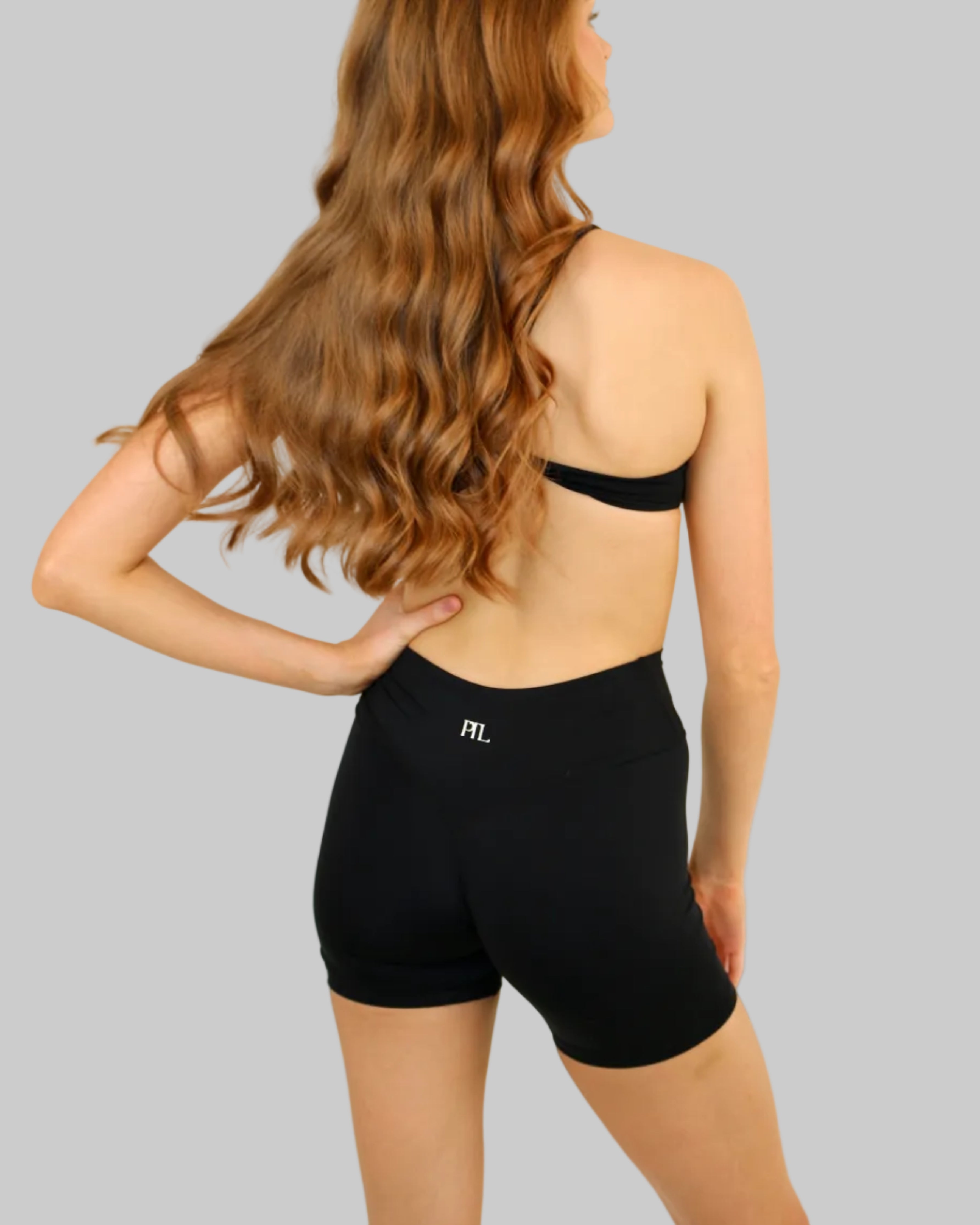 A woman with long wavy light brown hair is shown from behind wearing black athletic shorts and a black bandeau top against a plain off-white background.