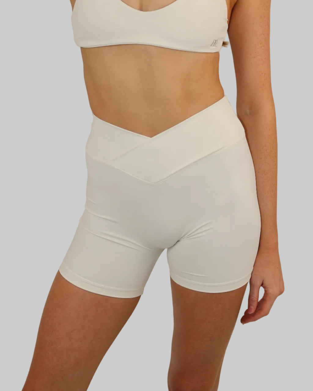 A model wearing a matching two-piece activewear set consisting of a white sports bra and high-waisted white cycling shorts with a V-cut waistband, set against a plain light beige background.